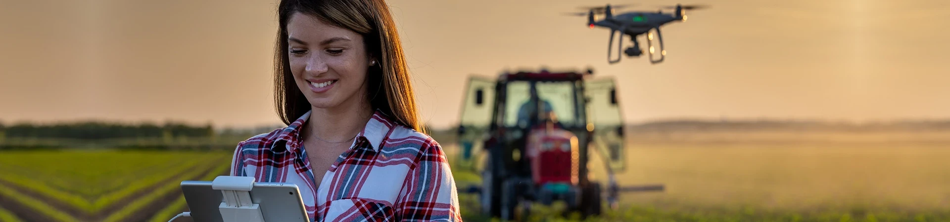 young woman driving drone with remote control in front of tractor in soybean field